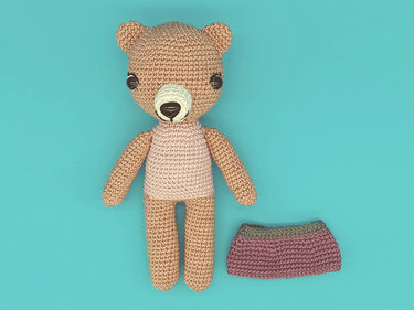 Gehäkelter Amigurumi-Teddy in Beige mit hellrosa Oberteil; daneben ein gehäkelter Rock in Altrosa auf türkisfarbenem Hintergrund