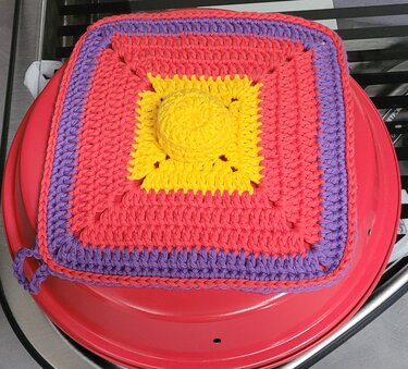 Quadratischer gehäkelter Topflappen mit gelbem Mittelkreis, korallroten Reihen, lila Rand und Aufhängeöse