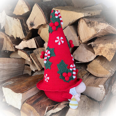 Gehäkelter Weihnachtswichtel mit großer roter, verzierter Mütze vor gestapeltem Kaminholz.