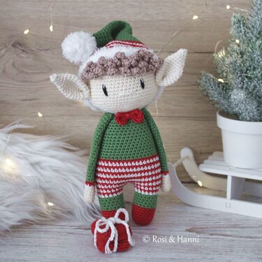 Gehäkelter Weihnachtself in Grün und Rot mit Zipfelmütze, roter Fliege und gestreifter Hose neben kleinem roten Geschenk vor weihnachtlicher Dekoration.