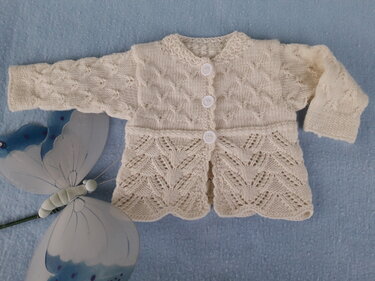 Cremefarbene gestrickte Babyjacke mit drei weißen Knöpfen, Zopf- und Ajourmuster auf blauem Untergrund
