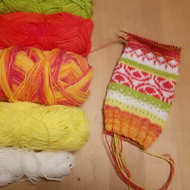 Teilweise gestrickte Socke mit buntem Jacquard- und Streifenmuster neben Garnknäueln auf einer Holzoberfläche.