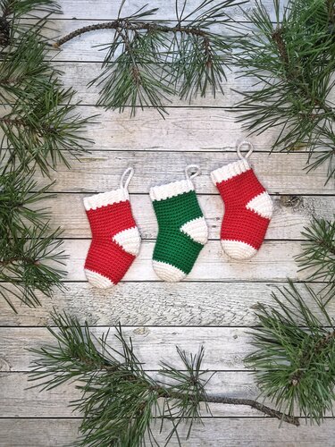 Three small crochet stocking ornaments in red and green with white trim and hanging loops on weathered wood with pine sprigs