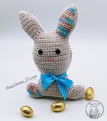 Beigefarbener gehäkelter Amigurumi-Osterhase mit blauer Schleife und goldenen Schokoeiern auf hellem Hintergrund