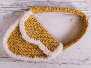Mustard yellow crocheted shoulder bag with soft white fluffy trim on a light wooden surface.