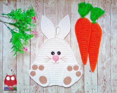 White crocheted bunny wall hanging with stitched paw pads and two orange crocheted carrots on a light wooden background