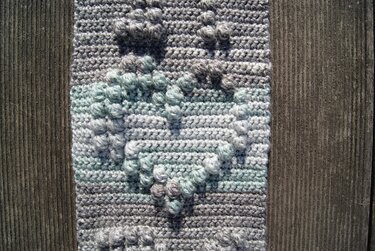 Nahaufnahme eines gehäkelten Stücks mit erhabenen Bobbelstichen in Grau, Weiß und Mint auf Holz.