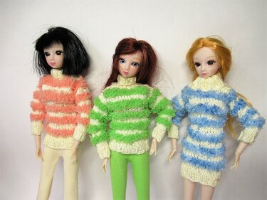 Drei Puppen in gestreiften, flauschigen Rollkragenpullover und einem Strickkleid in Koralle, Grün und Blau.