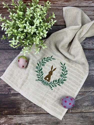 Beiges Handtuch mit besticktem braunen Hasen im grünen Blumenkranz, zwei Filz-Ostereier und kleine grüne Pflanze auf dunklem Holzuntergrund.