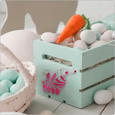 Mintgrüne Holzkiste mit pinkem Hasen- und Vogel-Plottmotiv, gefüllt mit pastellfarbenen Eiern und einer Filzkarotte