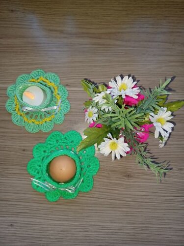 Zwei grüne gehäkelte Eierwärmer/Teelichthalter, einer mit einem braunen Ei, der andere mit einem Teelicht, daneben ein Blumenstrauß auf Holz.