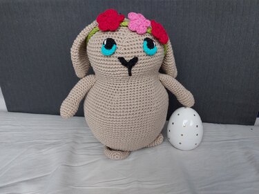 Beiger Amigurumi-Häkelhase mit türkisfarbenen Augen und rosa-rotem Blumenkranz neben weißem Ei mit goldenen Punkten.