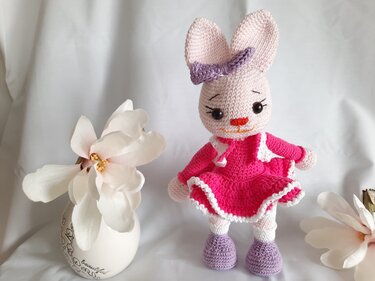 Gehäkelter Amigurumi-Hase in rosa Kleid mit lila Schleife neben Blüte