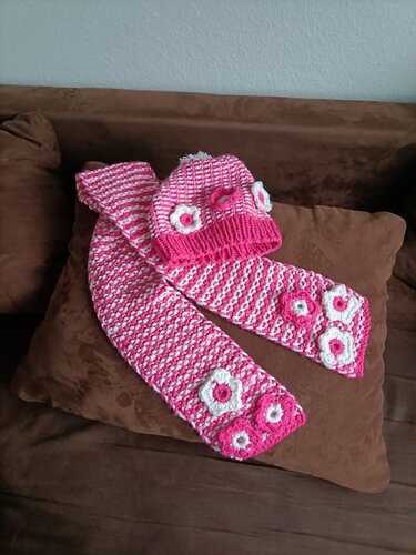 Gestreiftes Strickmützen- und Schal-Set in Pink und Weiß mit Blütenapplikationen, auf braunem Polster liegend
