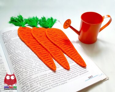 Three orange crocheted carrot bookmarks with green tops lying on an open book beside a miniature orange watering can.