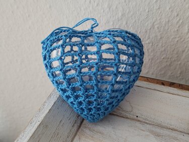 Herzförmiges Glasgefäß mit blauem, grobmaschigem Häkelnetz auf heller Holzfläche vor einer weißen Wand.