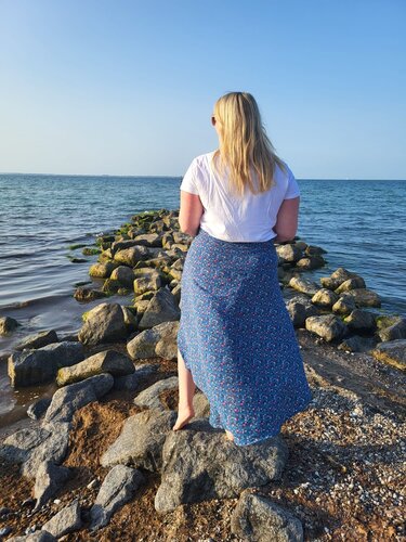 Rückenansicht einer Frau in weißem T‑Shirt und blauem Maxirock mit seitlichem Schlitz, barfuß auf Felsen vor dem Meer