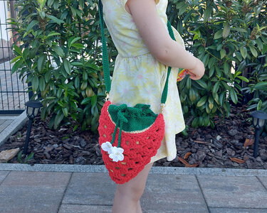 Kind trägt gehäkelte Erdbeer-Umhängetasche: rot mit grünem Zugband, grünem Schultergurt, goldfarbenen Karabinern und weißer Häkelblüte