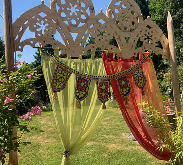 Gehäkelte Banner-Girlande mit bunten Mustern und Quasten an einer geschnitzten Holzpergola neben grünen und roten Vorhängen