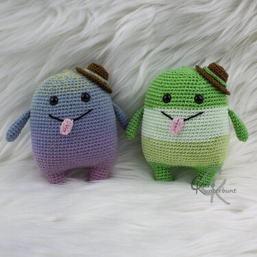 Zwei kompakte Amigurumi: links violett-blauer Farbverlauf, rechts grün-weiß-grün gestreift; beide mit kleinen braunen Häkelhüten, schwarzen Augen und rosa Zungen auf weißem Fellhintergrund.