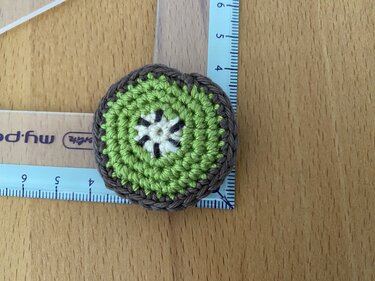 Mini gehäkelte Kiwi-Applikation in Grün, Braun und Weiß auf Holzuntergrund neben Lineal
