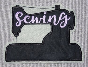 Gesticktes Nähmaschinenmotiv in Schwarz mit lila ‚Sewing‘-Schriftzug auf hellgrauem Grund