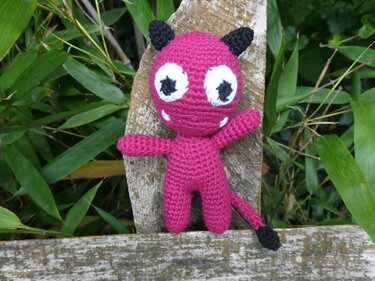 Gehäkeltes fuchsiafarbenes Amigurumi-Teufelchen mit schwarzen Hörnern und Schwanzspitze, großen Augen und weißen Fangzähnchen. Es sitzt auf einer verwitterten Holzbank vor grünen Pflanzen.