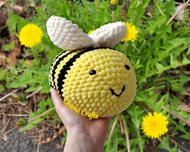 Hand holding a yellow and black crochet bee amigurumi with white wings over grass and dandelions