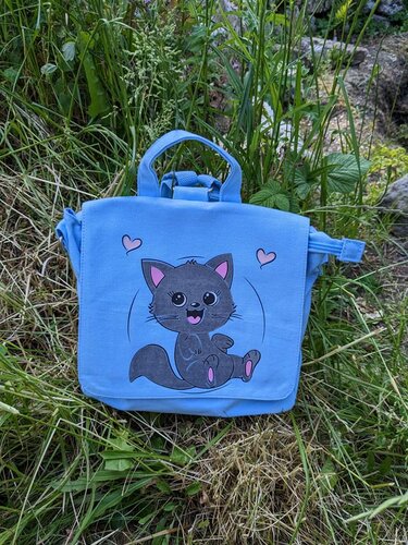 Hellblaue Stofftasche im Gras mit Plottmotiv einer grauen Katze und rosa Herzen
