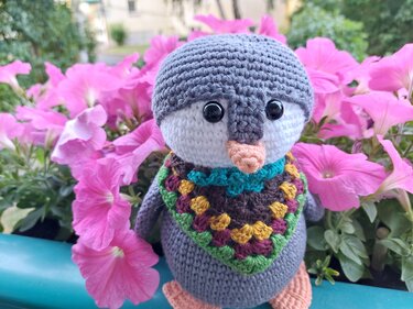 Gehäkelter Amigurumi-Pinguin mit großem Kopf, schwarzen Augen, orangefarbenem Schnabel und buntem Granny-Square-Schal vor pinken Petunien.