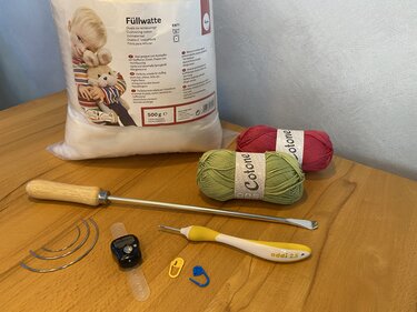 Häkelzubehör auf Holztisch: Packung Füllwatte, grüne und rosa Garnknäuel, Häkelnadel 2,5 mm, Reihenzähler, Maschenmarkierer und Stopfwerkzeuge.