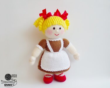 Hand-knitted girl doll in brown dress and white apron with yellow hair and red bows.
