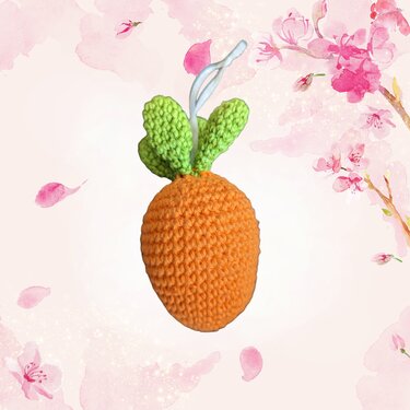Gehäkelte orange Karotte mit grünen Blättern und weißer Schlaufe vor zartem, blumigem Hintergrund.