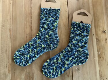 Zwei blau-gelb gemusterte Stricksocken auf hellem Holz mit braunen Etiketten.