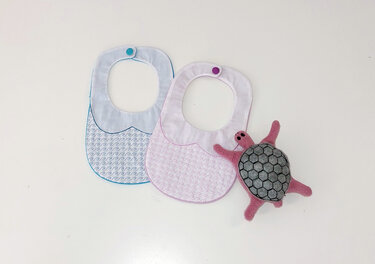 Zwei bestickte Baby-Lätzchen in Blau und Rosa mit Druckknopfverschluss neben einer gefilzten Schildkröte