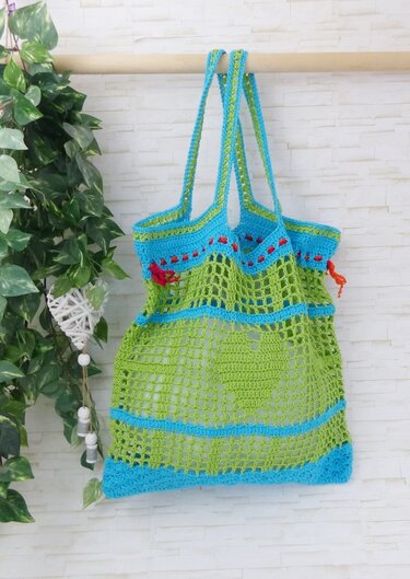 Handgehäkelte Henkeltasche in Grün und Blau mit roten Details, aufgehängt vor heller Wand neben Pflanze