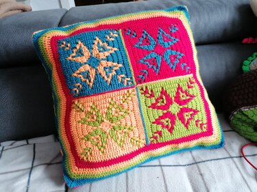 Gehäkeltes Kissen mit vier bunten Quadraten in Blau, Pink, Orange und Grün auf grauem Sofa