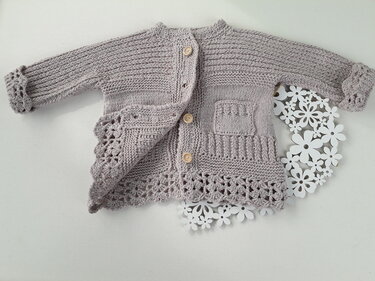 Hellgraues gestricktes Babyjäckchen mit runden Knöpfen, Rippen- und Lochmustern, aufgesetzter Tasche und gehäkelter Spitzenkante auf hellem Hintergrund mit Papierblumen
