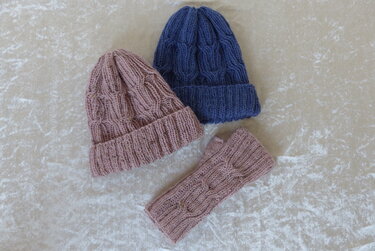 Zwei gefaltete gestrickte Beanies, eine rosa und eine blau, neben einer rosa Handstulpe auf hellem Stoff
