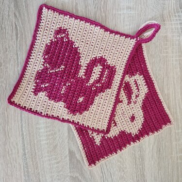 Zwei gehäkelte quadratische Topflappen mit Hasenmotiv in Pink und Beige auf hellem Holzboden.