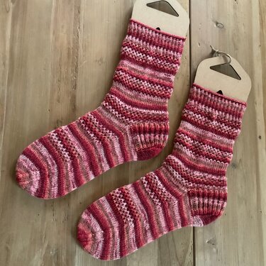 Handgestrickte Socken in Rot- und Rosatönen mit Streifen- und Lochmuster auf Holz liegend, mit Pappaufhängern