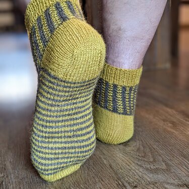 Handgestrickte Socken in Senfgelb und Dunkelgrau mit Streifenmuster, Nahaufnahme von Fuß und Ferse auf Holzboden.