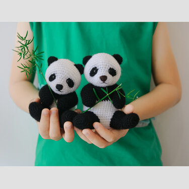 Pair of black-and-white crocheted panda amigurumi, each holding a small bamboo sprig, held in a person's hands.