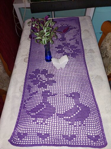 Violetter gehäkelter Tischläufer im Filetmuster auf hellem Tischtuch, mit blauer Vase und Zimmerpflanze