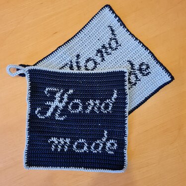Zwei quadratische gehäkelte Topflappen in Dunkelblau und Hellgrau mit Einfassung, 'Hand made' Schriftzug. Der dunkelblaue Topflappen verfügt über eine Aufhängeschlaufe.