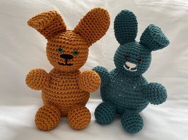 Zwei gehäkelte Amigurumi-Hasen in Orange und Petrol sitzen nebeneinander vor weißem Hintergrund.