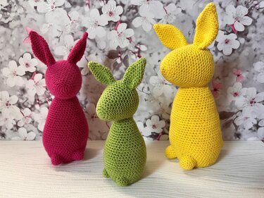 Drei gehäkelte Amigurumi-Hasen in Pink, Hellgrün und Gelb vor Blütenmuster-Hintergrund.