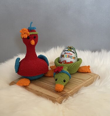 Zwei gehäkelte Amigurumi-Vögel in Rot und Grün, einer davon dient als Halterung für ein Schokoladenei auf einer Holzplatte