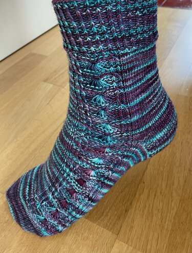 Handgestrickte Socke in Türkis, Lila und Weiß mit Noppen- und Zopfmuster.