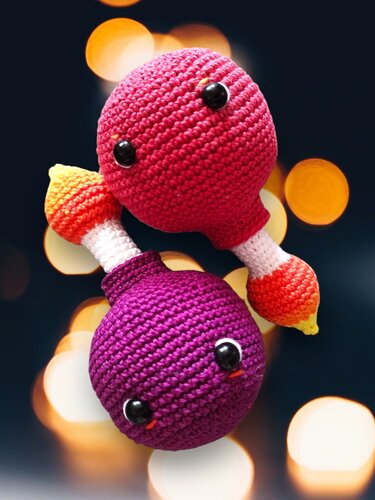 Zwei handgehäkelte Amigurumi-Rasseln, eine pink, eine lila, mit weißen Griffen und schwarzen Sicherheitsaugen